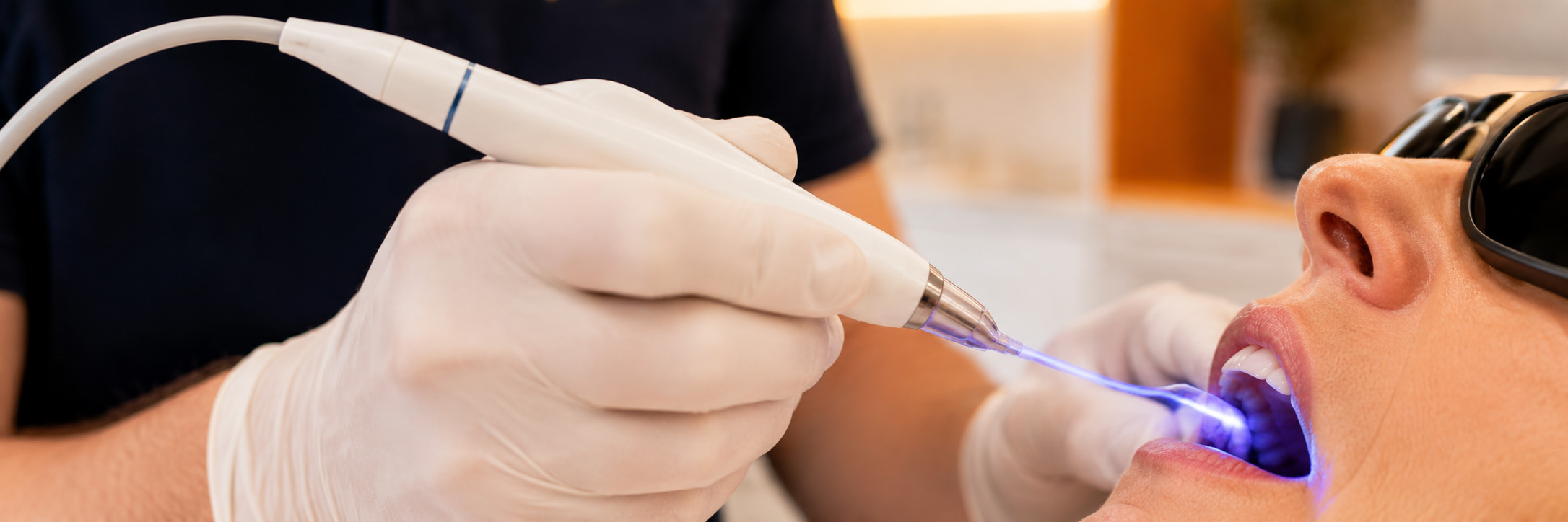 Ein Zahnarzt, der Handschuhe trägt, setzt im Mund eines Patienten ein zahnärztliches Lasergerät ein. Der Patient trägt während der zahnärztlichen Behandlung eine Schutzbrille.