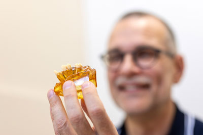 Ein Mann mit Brille und Schnurrbart hält ein gelbes Zahnmodell hoch und lächelt im Hintergrund, während das Modell im Vordergrund scharf abgebildet ist.