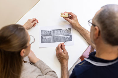 Zwei Personen betrachten einen Röntgenausdruck auf einem Tisch. Eine Person hält ein Zahnmodell in der Hand, während die andere auf das Röntgenbild zeigt. Beide tragen eine Brille und scheinen sich über Zahngesundheit zu unterhalten.