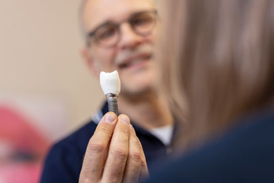 Eine Person hält ein Zahnimplantat hoch, während eine andere Person mit Brille im Hintergrund zuschaut und lächelt. Das Implantat hat eine weiße, zahnförmige Krone und einen Metallschraubfuß.