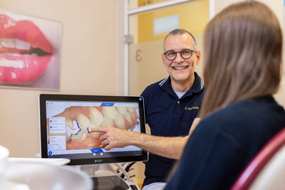 Ein Zahnarzt lächelt, während er einem Patienten in einem Untersuchungszimmer ein Zahnmodell auf einem Computerbildschirm zeigt. Im Hintergrund hängt ein Poster mit Lippen an der Wand.