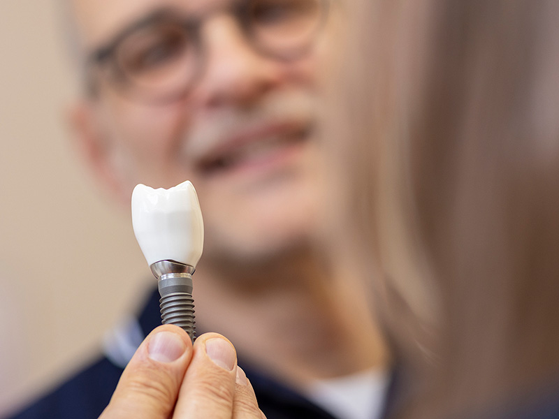 implantatberatung Im Vordergrund hält eine Person ein Zahnimplantat mit einer Zahnkrone in der Hand, während im Hintergrund ein unscharf abgebildeter Mann mit Brille und Schnurrbart lächelt.